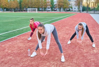 Futbol stadyumunda esneme egzersizleri yapan üç kız.