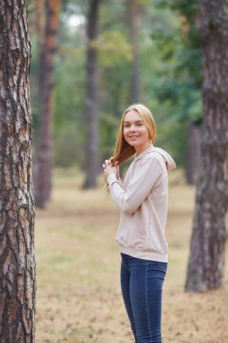 Bej kapüşonlu güzel sarışın kadın parkta yürüyor. Çekici bir kadın parkta aktif zaman geçiriyor. Sonbahar parkında eğlenen neşeli genç bir kadının portresi. Doğada rahatla..
