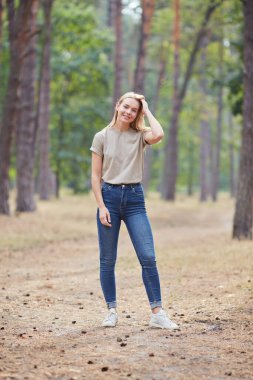 Çam ormanında poz veren doğal makyajlı sarışın kadın. Güzel mavi gözlü, bej tişörtlü beyaz kadın yaz tatilinin tadını çıkarıyor. Vahşi yaşam ve insanlar kavramı.