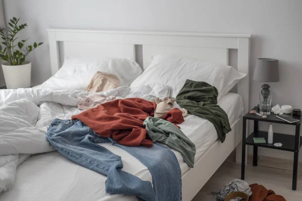 服はベッドの周りに投げられた 白い寝具とミニマリズムのモダンなインテリアのクラッタリングベッドルーム — ストック写真