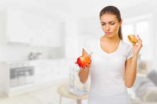 Chica dudando con manzana y hamburguesa en la sala de luz —  Fotos de Stock