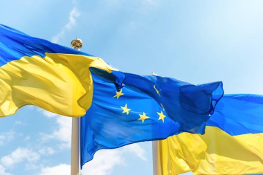 Ukraine and European Union flags on poles fluttering by wind on blue sky. Ukrainian flag