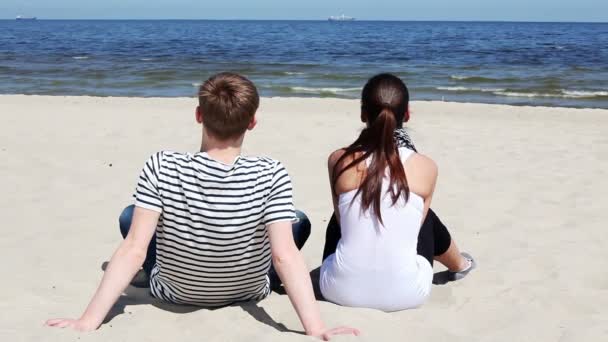 Молодая пара сидит на песке на пляже в повседневной одежде и смотрит на волны — стоковое видео