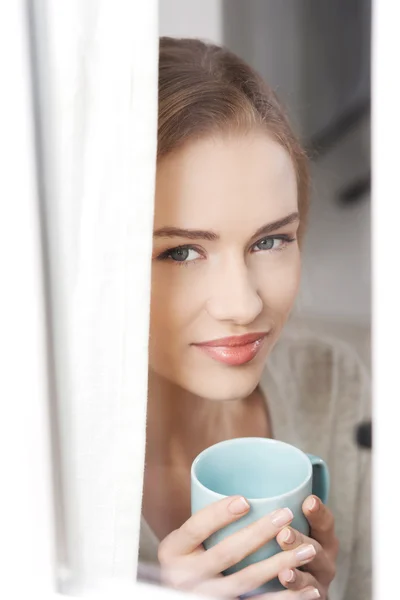 Jeune belle femme tenant une tasse et regardant par la fenêtre . — Photo