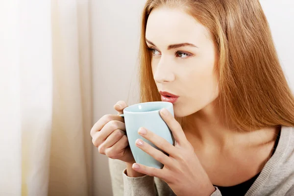 Giovane bella donna in possesso di una tazza e guardando attraverso la finestra . — Foto Stock