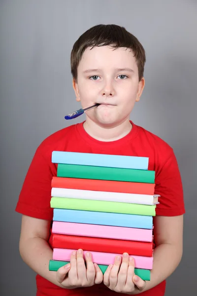 Niño con un bolígrafo en la boca sosteniendo una pila de libros — Foto de Stock