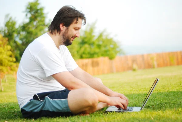 El hombre joven está acostado en un terreno verde con el ordenador portátil y tomando un buen tiempo —  Fotos de Stock