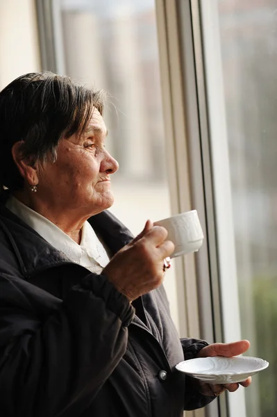 Retrato de una mujer mayor mirando por la ventana y bebiendo una taza de café —  Fotos de Stock