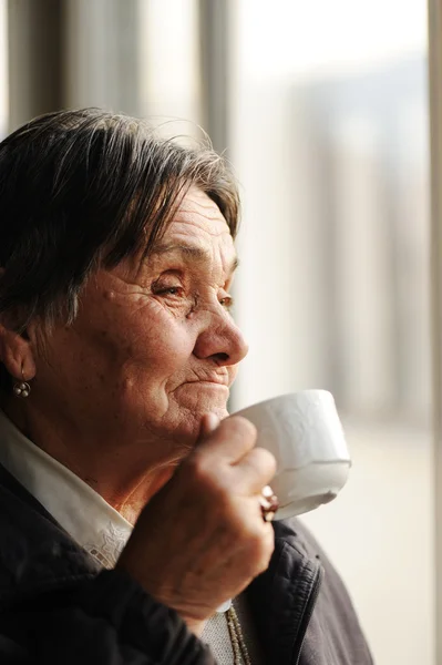Porträtt av äldre kvinna tittar genom fönstret och dricka kaffe — Stockfoto