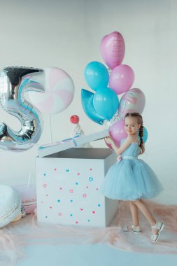 Mavi elbiseli sevimli, gülümseyen küçük kız. İyi ki doğdun Doğum günü prensesi bir balon zeminine karşı pastayla. Hediyelik eşya dükkanı. Alışveriş günü. Çocukluk ve komik kutlama konsepti.