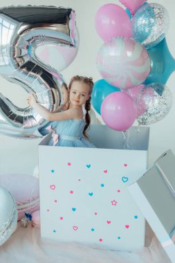 Mavi elbiseli sevimli, gülümseyen küçük kız. İyi ki doğdun Doğum günü prensesi bir balon zeminine karşı pastayla. Hediyelik eşya dükkanı. Alışveriş günü. Çocukluk ve komik kutlama konsepti.