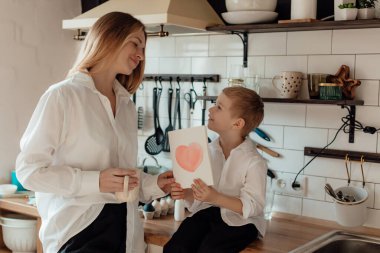 Mutlu küçük çocuk gülümseyen anneyi tebrik ediyor ve evde tatil kutlamalarında kırmızı kalple kart veriyor. Aile hayatı kutlamaları, Anneler Günü konsepti kutlandı.