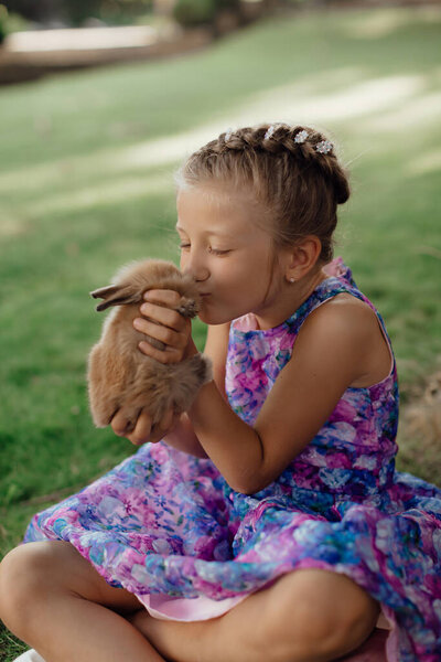 Little girl sitting on the green grass with rabbit. Cute child girl holding a bunny in her hands on Easter day. Fluffy brown plush pet.