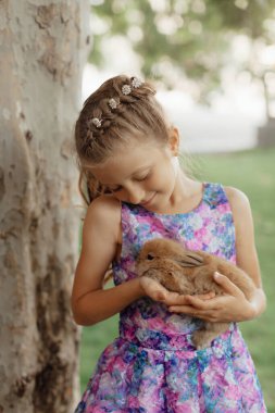 Küçük kız elinde tavşanla yeşil çimlerde oturuyor. Paskalya günü elinde tavşan tutan tatlı bir kız çocuğu. Yumuşak kahverengi peluş evcil hayvan.