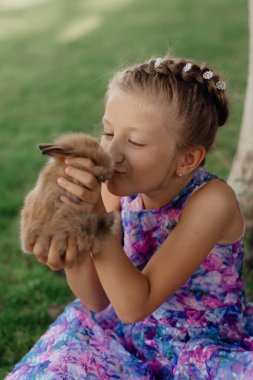 Küçük kız elinde tavşanla yeşil çimlerde oturuyor. Paskalya günü elinde tavşan tutan tatlı bir kız çocuğu. Yumuşak kahverengi peluş evcil hayvan.