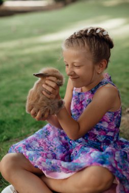 Küçük kız elinde tavşanla yeşil çimlerde oturuyor. Paskalya günü elinde tavşan tutan tatlı bir kız çocuğu. Yumuşak kahverengi peluş evcil hayvan.