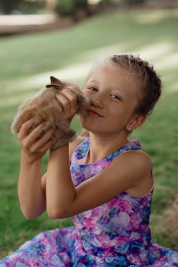Küçük kız elinde tavşanla yeşil çimlerde oturuyor. Paskalya günü elinde tavşan tutan tatlı bir kız çocuğu. Yumuşak kahverengi peluş evcil hayvan.