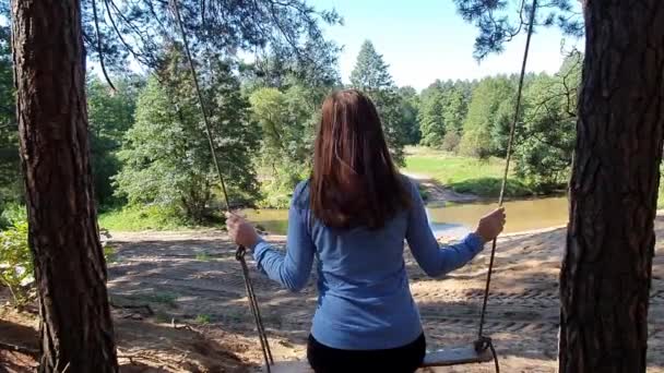 Молодая красивая женщина, качающаяся на деревянных качелях, висящих на веревке на ветке дерева — стоковое видео