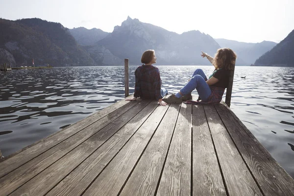 两个少女笑着坐在湖边的一个码头上和后面的群山上 — 图库照片