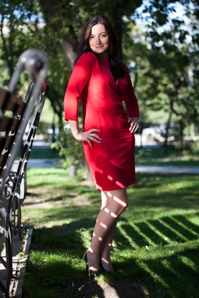 Girl in the red dress is standing in a park near the bench — Stock Photo, Image