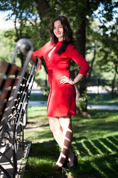 Girl in the red dress is standing in a park near the bench — Stock Photo, Image