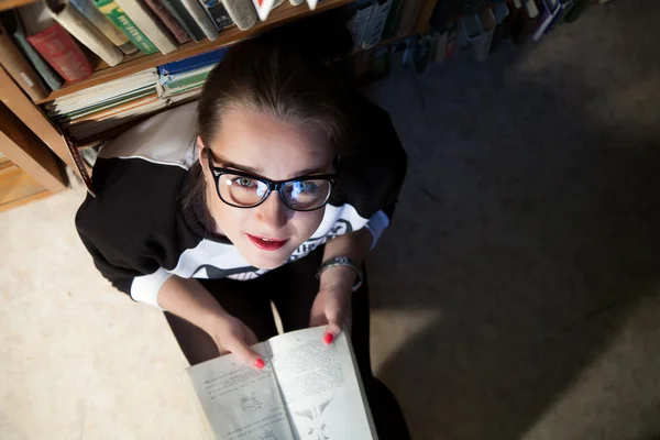 Young girl with glasses sitting in a rack with books — Stock Photo, Image