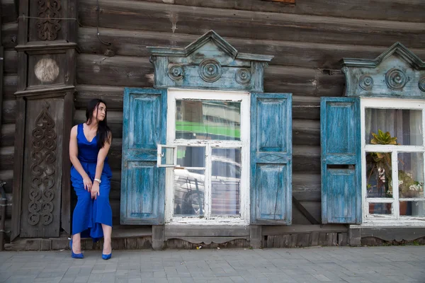 Menina em um vestido azul em um fundo de uma velha casa de madeira — Fotografia de Stock