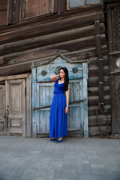 Meisje in een blauwe jurk op een achtergrond van een oud houten huis — Stockfoto