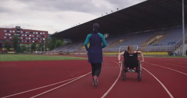 Žena s postižením jezdí na invalidním vozíku na atletické tréninkové dráze s muslimskou ženou na sobě hidžáb sportovní oblečení — Stock video