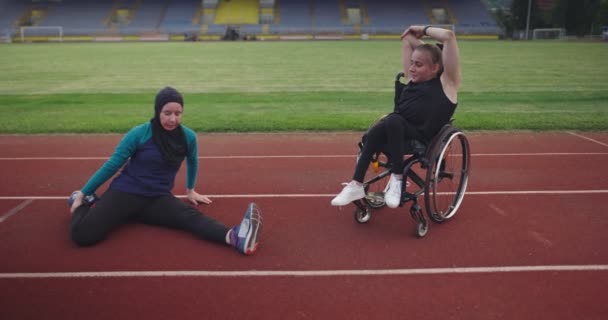 Kobieta z niepełnosprawnością rozgrzewa się na wózku inwalidzkim na torze treningowym lekkoatletycznym z muzułmanką ubraną w strój sportowy hidżab — Wideo stockowe