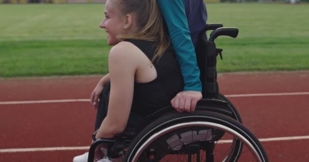 Una mujer con discapacidades montando una silla de ruedas en una pista de entrenamiento de atletismo con una mujer musulmana usando ropa deportiva de hijab — Vídeos de Stock