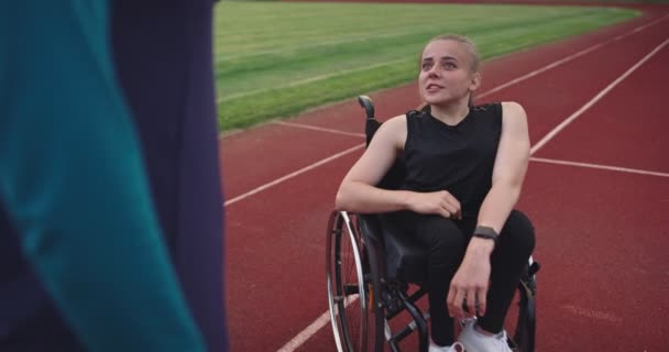 Eine behinderte Frau wärmt sich im Rollstuhl auf der Leichtathletik-Trainingsstrecke mit muslimischer Frau in Hijab-Sportbekleidung auf — Stockvideo