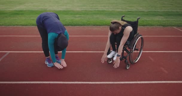 Kobieta z niepełnosprawnością rozgrzewa się na wózku inwalidzkim na torze treningowym lekkoatletycznym z muzułmanką ubraną w strój sportowy hidżab — Wideo stockowe