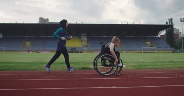 Eine behinderte Frau im Rollstuhl auf einer Leichtathletik-Trainingsstrecke mit muslimischer Frau in Hijab-Sportbekleidung — Stockvideo