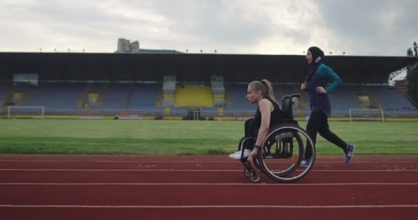 Una donna con disabilità guida una sedia a rotelle su una pista di atletica con donna musulmana che indossa abbigliamento sportivo hijab — Video Stock