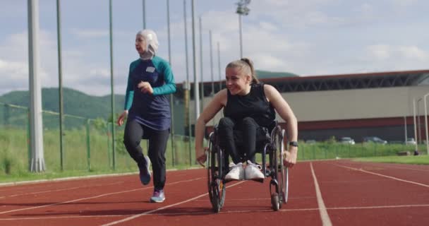 Kobieta z niepełnosprawnością na wózku inwalidzkim na torze treningowym z muzułmanką ubraną w strój sportowy hidżab — Wideo stockowe