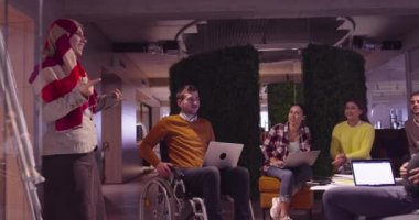 Modern bir açık alanda çalışan Müslüman bir kadın takıma bir sunum yapıyor. Orta Doğulu Arap iş kadını yaratıcı bir ofiste. Ekip toplantısında tekerlekli sandalyedeki engelli iş adamı 