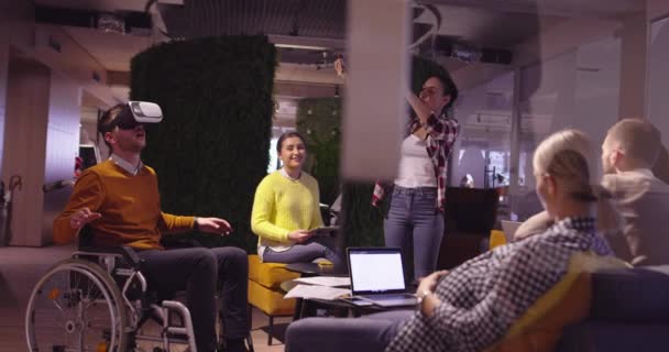 Disabled businessman in a wheelchair at work in modern open space coworking office on team meeting using virtual reality goggles and drone simulation. Effective teamwork in a startup business. — Stock Video