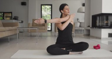 Spor giyim sektöründe karışık bir yarış kadını dizüstü bilgisayarda yoga eğitimi alırken esneme egzersizleri yapıyor. Arka planda kanepe ile oturma odasında egzersiz yapan sağlıklı bir kız..