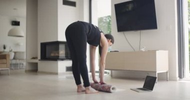 Spor giyim sektöründe karışık bir yarış kadını dizüstü bilgisayarda yoga eğitimi alırken esneme egzersizleri yapıyor. Arka planda kanepe ile oturma odasında egzersiz yapan sağlıklı bir kız..