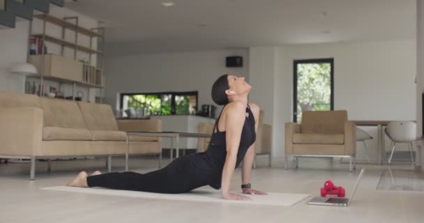 身穿运动服的混血女运动员一边做伸展运动 一边在线观看笔记本电脑上的瑜伽训练课 健康的女孩在客厅里锻炼 背靠沙发 — 图库视频影像