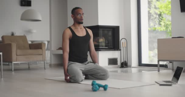 Afro-Amerikan atletik adam dizüstü bilgisayarda online eğitim dersleri izliyor, eğitmenden sonra hareketleri tekrarlıyor, evde bacak egzersizleri yapıyor. Kendini soyutlama. Karantina. — Stok video
