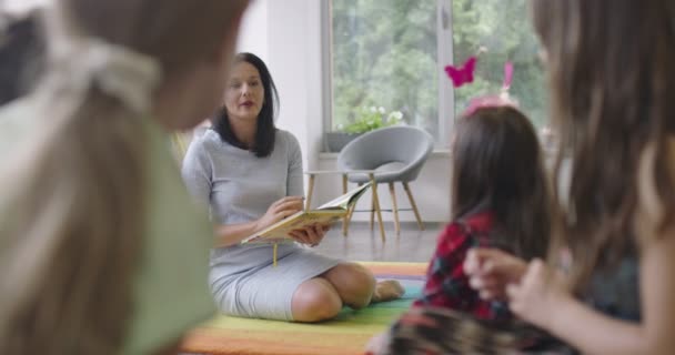 Storytime elementary or primary school teacher reading a story to a group of children in a daycare center. — Stockvideo