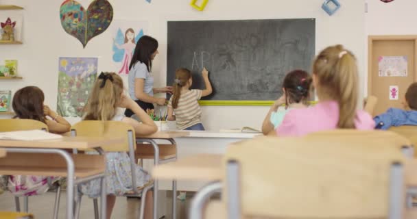 Escuela Primaria. La maestra ayudando al niño estudiante mientras escribe la respuesta en la pizarra. — Vídeo de stock