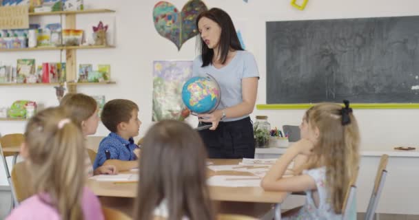 Kvinnlig lärare med barn i geografi klass tittar på jordklotet. Sidovy över grupp av olika glada skolbarn med världen i klassrummet i skolan. — Stockvideo