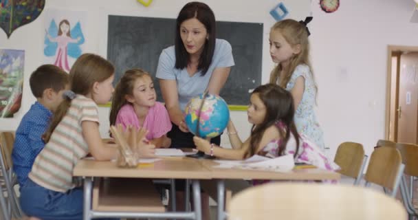 Kvinnlig lärare med barn i geografi klass tittar på jordklotet. Sidovy över grupp av olika glada skolbarn med världen i klassrummet i skolan. — Stockvideo