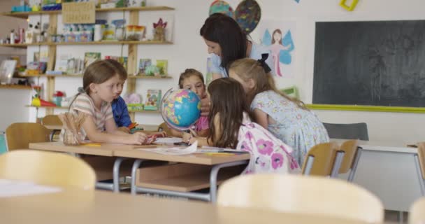 Női tanár gyerekekkel földrajz órán, akik a földgömböt nézik. Oldalnézet csoport változatos boldog iskolás gyerekek gömb osztályteremben az iskolában. — Stock videók