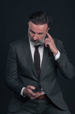 Müşteriyle sohbet et. Takım elbiseli yakışıklı genç adam, akıllı telefonunu kullanıyor ve koyu gri arka planda dikilirken gülümsüyor. Yüksek kalite fotoğraf