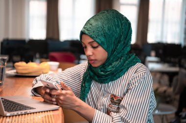 Afrikalı Müslüman iş kadını yeşil başörtüsü takıyor. Modern açık plan başlangıç ofisindeki rahatlama alanında dizüstü bilgisayarda çalışırken akıllı telefon kullanıyor. Yüksek kaliteli fotoğraf.