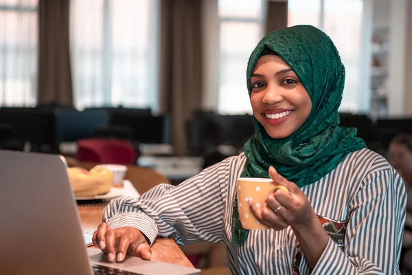 アフリカ系イスラム教徒のビジネス女性は 現代のオープンプランのスタートアップオフィスでノートパソコンのリラクゼーションエリアで働いている間 緑茶ヒジャーブ飲料茶を身に着けています 高品質の写真 — ストック写真